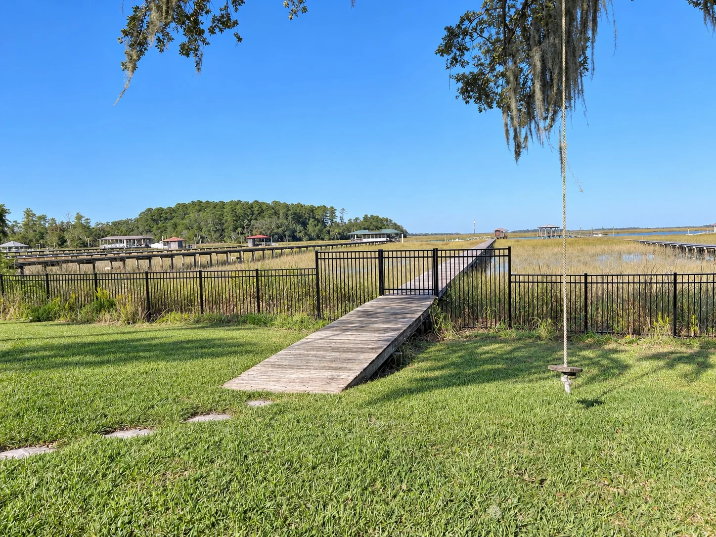 Yard view of a black aluminum picket fence and walk-through gate onto a wooden dock boardwalk, with the marsh behind, at a home on Whitemarsh Island in Savannah, Georgia